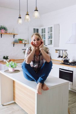Genç, sarışın, 30 'lu yaşlarda, kot pantolonlu ve temiz mutfağında oturan ve rahatlatıcı gömlekli bir kadın. Sağlıklı ve mutlu bir yaşam tarzı. Boş zaman.