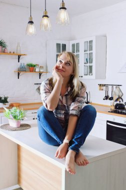 Genç, sarışın, 30 'lu yaşlarda, kot pantolonlu ve temiz mutfağında oturan ve rahatlatıcı gömlekli bir kadın. Sağlıklı ve mutlu bir yaşam tarzı. Boş zaman.