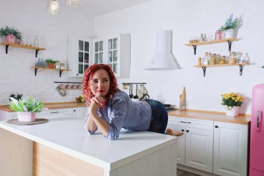 Genç, kızıl saçlı, kıvırcık saçlı, 30 'lu yaşlarda kot pantolonlu ve temiz mutfağında oturan ve rahatlatıcı gömleği olan bir kadın. Sağlıklı ve mutlu bir yaşam tarzı. Boş zaman. Ev hanımı.. 