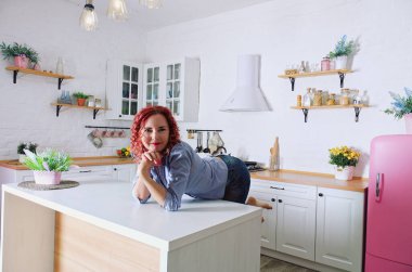 Genç, kızıl saçlı, kıvırcık saçlı, 30 'lu yaşlarda kot pantolonlu ve temiz mutfağında oturan ve rahatlatıcı gömleği olan bir kadın. Sağlıklı ve mutlu bir yaşam tarzı. Boş zaman. Ev hanımı.. 