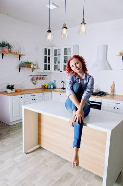 Genç, kızıl saçlı, kıvırcık saçlı, 30 'lu yaşlarda kot pantolonlu ve temiz mutfağında oturan ve rahatlatıcı gömleği olan bir kadın. Sağlıklı ve mutlu bir yaşam tarzı. Boş zaman. Ev hanımı.. 