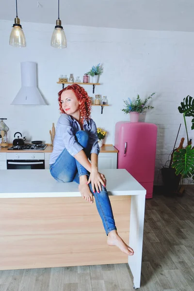 Genç, kızıl saçlı, kıvırcık saçlı, 30 'lu yaşlarda kot pantolonlu ve temiz mutfağında oturan ve rahatlatıcı gömleği olan bir kadın. Sağlıklı ve mutlu bir yaşam tarzı. Boş zaman. Ev hanımı.. 
