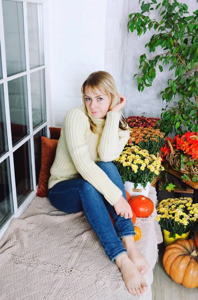 黄色のセーターの若いかわいいブロンドの女性は 秋のお祝いの装飾 オレンジカボチャ 秋の花と居心地の良い家の窓の席に座っています ティーカップ — ストック写真