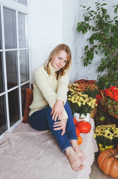 黄色のセーターの若いかわいいブロンドの女性は 秋のお祝いの装飾 オレンジカボチャ 秋の花と居心地の良い家の窓の席に座っています ティーカップ — ストック写真