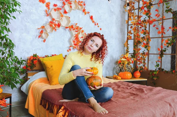 暖かいセーターを着た美しい赤毛の巻き若い女性が居心地の良い家でベッドの中に座って 笑って 楽しんでいます 秋の装飾 オレンジの葉 カボチャの家のインテリア — ストック写真