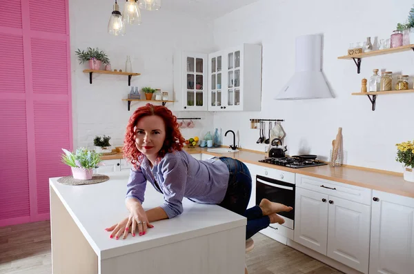 Genç, kızıl saçlı, kıvırcık saçlı, 30 'lu yaşlarda kot pantolonlu ve temiz mutfağında oturan ve rahatlatıcı gömleği olan bir kadın. Sağlıklı ve mutlu bir yaşam tarzı. Boş zaman. Ev hanımı.. 