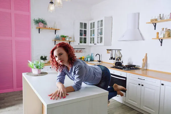 Genç, kızıl saçlı, kıvırcık saçlı, 30 'lu yaşlarda kot pantolonlu ve temiz mutfağında oturan ve rahatlatıcı gömleği olan bir kadın. Sağlıklı ve mutlu bir yaşam tarzı. Boş zaman. Ev hanımı.. 