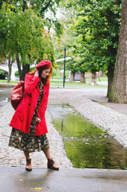 Kızıl saçlı, çilli, kırmızı bereli mavi gözlü, sonbahar parkında yürüyen, gülümseyen genç ve mutlu bir kadın. Sarı sonbahar yaprakları.