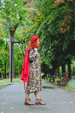 Kızıl saçlı, çilli, kırmızı bereli mavi gözlü, sonbahar parkında yürüyen, gülümseyen genç ve mutlu bir kadın. Sarı sonbahar yaprakları.