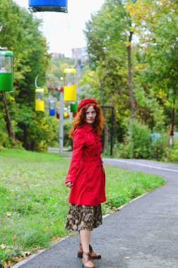 Kızıl saçlı, çilli, kırmızı bereli mavi gözlü, sonbahar parkında yürüyen, gülümseyen genç ve mutlu bir kadın. Sarı sonbahar yaprakları.