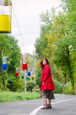 Kızıl saçlı, çilli, kırmızı bereli mavi gözlü, sonbahar parkında yürüyen, gülümseyen genç ve mutlu bir kadın. Sarı sonbahar yaprakları.