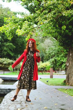 Genç, mutlu bir kadın, kızıl saçlı, çilli, kırmızı bereli mavi gözlü, sonbahar parkında yürüyen, kahve içen, gülümseyen. Sarı sonbahar sezonu.