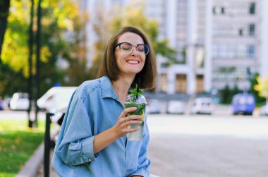 Gözlüklü ve günlük kıyafetleri olan zarif iş kadınının şehir portresi. Şehir caddesinde yürüyen kadın. Sağlıklı meyve suyu içmek, meyve suyu içmek, kitap okumak ve gün içinde dinlenmek.. 