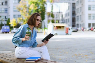 Gözlüklü ve günlük kıyafetleri olan zarif iş kadınının şehir portresi. Şehir caddesinde yürüyen kadın. Sağlıklı meyve suyu içmek, meyve suyu içmek, kitap okumak ve gün içinde dinlenmek.. 