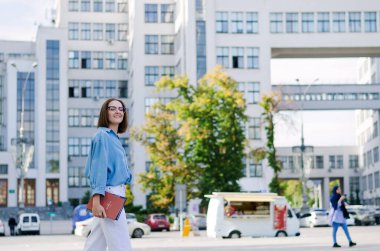 Şehirde gözlüklü ve günlük kıyafetleri olan zarif bir iş kadınının şehir portresi. Yeşil parkta yürüyen, güneşli bir günde rahatlayan, gülümseyen bir kadın. Hobi ve eğlence..