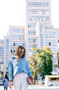 Şehirde gözlüklü ve günlük kıyafetleri olan zarif bir iş kadınının şehir portresi. Yeşil parkta yürüyen, güneşli bir günde rahatlayan, gülümseyen bir kadın. Hobi ve eğlence..