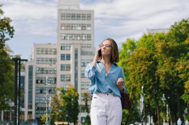 Gözlüklü ve günlük kıyafetleri olan zarif iş kadınının şehir portresi. Şehir caddesinde çeşmenin yanında yürüyen, telefonuyla konuşan, gündüzleri şehir caddesinde çalışan bir kadın.