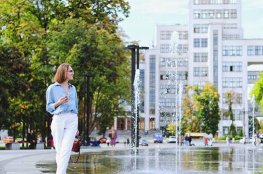 Gözlüklü ve günlük kıyafetleri olan zarif iş kadınının şehir portresi. Şehir caddesinde çeşmenin yanında yürüyen, telefonuyla konuşan, gündüzleri şehir caddesinde çalışan bir kadın.