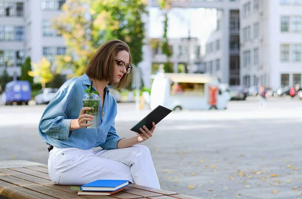 Gözlüklü ve günlük kıyafetleri olan zarif iş kadınının şehir portresi. Şehir caddesinde yürüyen kadın. Sağlıklı meyve suyu içmek, meyve suyu içmek, kitap okumak ve gün içinde dinlenmek.. 