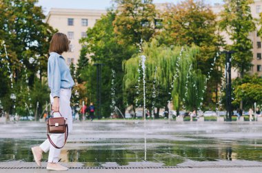 Şehirde gözlüklü ve günlük kıyafetleri olan zarif bir iş kadınının şehir portresi. Yeşil parkta yürüyen, güneşli bir günde rahatlayan, gülümseyen bir kadın. Hobi ve eğlence..