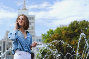 Gözlüklü ve günlük kıyafetleri olan zarif iş kadınının şehir portresi. Şehir caddesinde çeşmenin yanında yürüyen, telefonuyla konuşan, gündüzleri şehir caddesinde çalışan bir kadın.