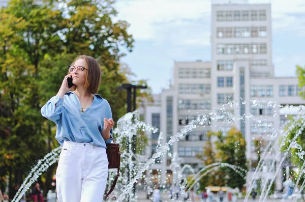 Gözlüklü ve günlük kıyafetleri olan zarif iş kadınının şehir portresi. Şehir caddesinde çeşmenin yanında yürüyen, telefonuyla konuşan, gündüzleri şehir caddesinde çalışan bir kadın.