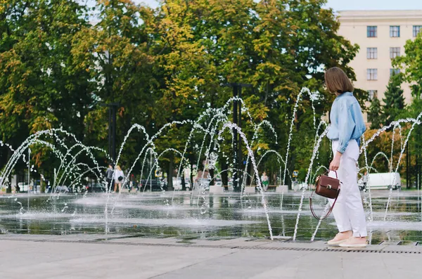 Şehirde gözlüklü ve günlük kıyafetleri olan zarif bir iş kadınının şehir portresi. Yeşil parkta yürüyen, güneşli bir günde rahatlayan, gülümseyen bir kadın. Hobi ve eğlence..