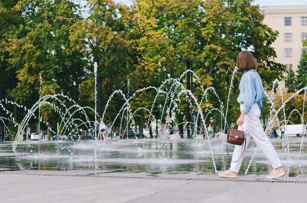 Şehirde gözlüklü ve günlük kıyafetleri olan zarif bir iş kadınının şehir portresi. Yeşil parkta yürüyen, güneşli bir günde rahatlayan, gülümseyen bir kadın. Hobi ve eğlence..