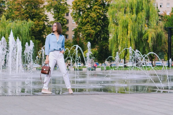 Şehirde gözlüklü ve günlük kıyafetleri olan zarif bir iş kadınının şehir portresi. Yeşil parkta yürüyen, güneşli bir günde rahatlayan, gülümseyen bir kadın. Hobi ve eğlence..