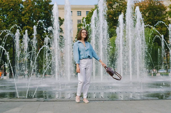 Şehirde gözlüklü ve günlük kıyafetleri olan zarif bir iş kadınının şehir portresi. Yeşil parkta yürüyen, güneşli bir günde rahatlayan, gülümseyen bir kadın. Hobi ve eğlence..