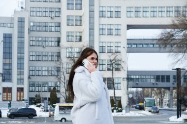 Kışın günlük giysiler içinde, beyaz ceketli zarif bir iş kadınının şehir portresi. Şehir caddesinde yürümek, telefonda konuşmak, selfie çekmek. Açık havada çalışmak.