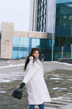 Kışın günlük giysiler içinde, beyaz ceketli zarif bir iş kadınının şehir portresi. Şehir caddesinde yürümek, telefonda konuşmak, selfie çekmek. Açık havada çalışmak.