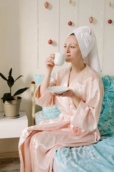 快適な家でパジャマのベッドに座っている彼女の頭の上にタオルを持つ若い幸せな女性 朝のコーヒーを飲む 美人ルーチン — ストック写真