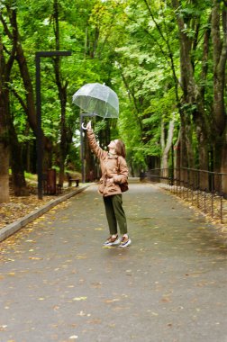 Yeşil parkta şemsiyeyle yağmurda yürüyen, gülümseyen, eğlenceli genç bir kadın. Bankta oturan kişi. Soğuk hava. Bahar. Düş.