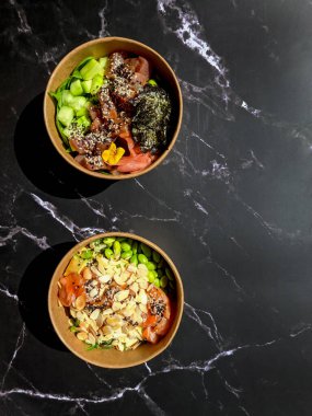 Tava kutusunun üst görüntüsü, siyah arka planda bir kase pirinç. Somon, avokado, soğan, mantar, brokoli ve susamlı pilav. Çin, Asya yemeği teslimatı. Tavalar için ambalaj. 