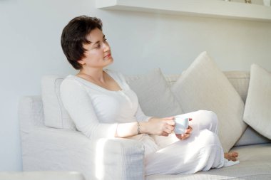 Beyaz etekli, rahat gömlekli, rahat rahat kanepede oturan mutlu bir kadının hayat tarzı portresi. Kahve içiyorum. Hayal kurmak.