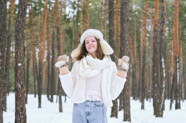 funny young woman in jeans, white hat and jacket in snowy park, forest. Girl walking near snow covered pine trees.  Having fun. Family winter holidays.