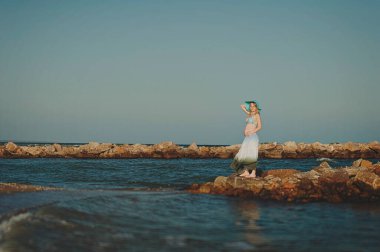 Beautiful young elegant pregnant blond woman in a blue hat walks on the beach, seashore at sunset. Azov sea. Motherhood.