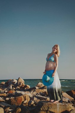 Beautiful young elegant pregnant blond woman in a blue hat walks on the beach, seashore at sunset. Azov sea. Motherhood.