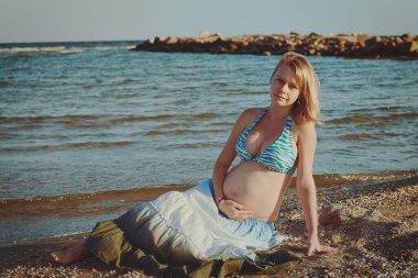 Beautiful young elegant pregnant blond woman in a blue hat walks on the beach, seashore at sunset. Azov sea. Motherhood.