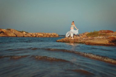 Beautiful young elegant pregnant blond woman in a blue hat walks on the beach, seashore at sunset. Azov sea. Motherhood.
