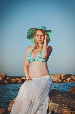 Beautiful young elegant pregnant blond woman in a blue hat walks on the beach, seashore at sunset. Azov sea. Motherhood.
