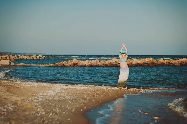 Beautiful young elegant pregnant blond woman in a blue hat walks on the beach, seashore at sunset. Azov sea. Motherhood.