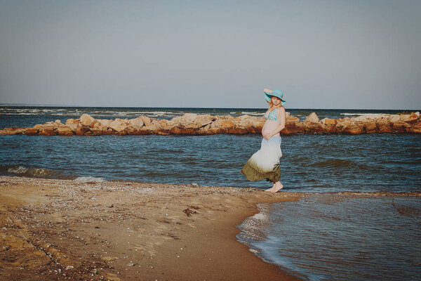 Beautiful young elegant pregnant blond woman in a blue hat walks on the beach, seashore at sunset. Azov sea. Motherhood.