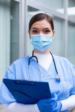 Kadın bir doktorun belinden yakın plan portresi, genç bir NHS İngiliz kadın Kafkas tıp uzmanı koruyucu yüz maskesi, eldiven, stetoskop, hasta dosyasını tutuyor.