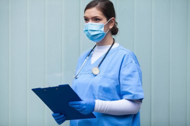 Mavi cam duvarın önünde dikilmiş mavi koruyucu ekipman, maske, eldiven, stetoskop, elinde dosya olan beyaz saçlı güzel bir kadının portresi.