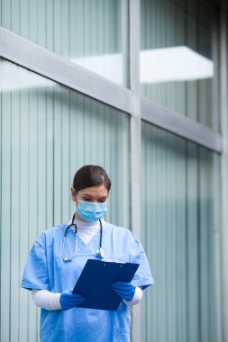 Hasta panosu formunu tutan kadın doktor hastane koridorundaki verileri analiz ediyor, mavi koruyucu PPE önlükler giyiyor: stetoskop, cerrahi koruyucu yüz maskesi, eldivenler. COVID-19 salgın krizi