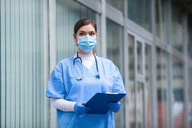 Sağlık merkezinin dışında duran, mavi üniformalı, koruyucu cerrahi maske takan, mavi eldivenli ve steteskoplu kadın bir NHS doktoru hasta dosyasını tutuyor, dalgın dalgın bakıyor.