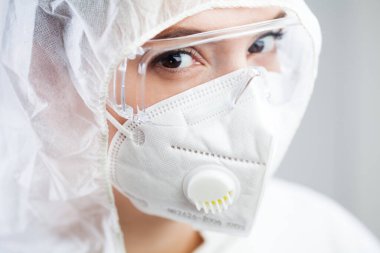 Güzel beyaz kadın tıbbi laboratuvar çalışanı portresi, korku dolu gözlere yakın, koruyucu gözlük, tulum, N95 yüz maskesi takan, kameraya bakan, beyaz arka planda izole edilmiş.