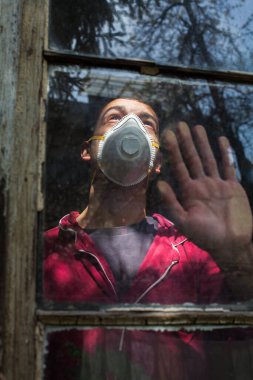 Koruyucu N95 yüz maskesi takan genç adam, camdan dışarı bakıyor, eli camın üzerinde, COVID-19 karantinasında depresif ve endişeli, Coronavirus 'a karşı güvenlik önlemi yayılıyor.